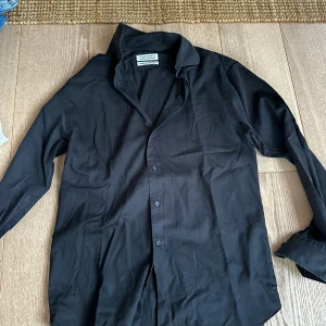 Svart skjorta från The Shirt Factory - Snygg svart skjorta från The Shirt Factory, designad av Linnéa Braun. Skjortan har klassisk krage, långa ärmar och knappar framtill. Skräddarsydd passform och tillverkad i mjuk bomull som är enkel att tvätta och bära. Perfekt för dig som gillar stilrena plagg.