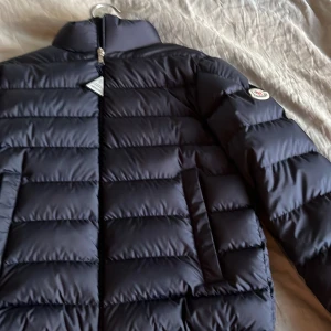 Svart Moncler pufferjacka herr - Snygg svart pufferjacka från Moncler med klassisk quiltad design och hög krage. Jackan har dragkedja framtill, två sidofickor och ikonisk Moncler-logga på ärmen. Materialet är glansigt och känns lyxigt, perfekt för kalla dagar.