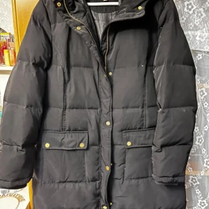 Svart lång pufferjacka med fuskpäls - Svart lång pufferjacka med huva och avtagbar fuskpäls. Jackan har guldiga knappar, dragkedja och stora fickor framtill. Perfekt för kalla dagar och har en klassisk, stilren look som funkar till det mesta. Har används två gånger men suttit i garderoben för fyra år. 