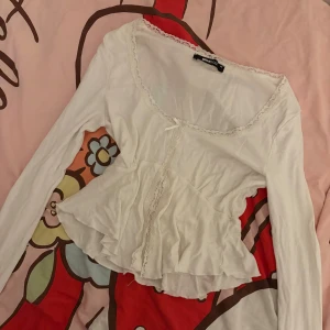 Vit spetsdekor blus från Gina Tricot - Supersöt vit blus från Gina Tricot med långa ärmar, spetsdetaljer längs urringningen och framtill samt en liten rosett vid halsen. Blusen har en lätt volang nertill och en feminin, romantisk vibe. Perfekt för dig som gillar en söt och trendig look.