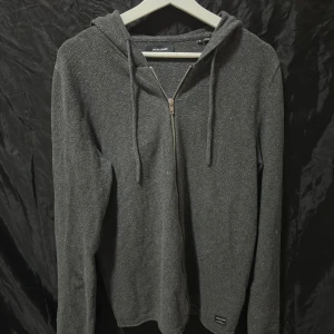 Mörkgrå hoodie från Jack & Jones - Mörkgrå hoodie från Jack & Jones med dragkedja framtill och huva med snörning. Tröjan är långärmad och har en enkel, stilren design i mjukt material. Perfekt för chill dagar eller när du vill ha något bekvämt och snyggt. Säljer den för att den är lite smått stor på mig och inte kommer till så stor användning. Storlek M skick 8,5/10