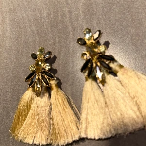 Guldiga örhängen med tofsar och stenar - Unika örhängen med guldiga detaljer, glittrande stenar och stora ljusbeiga tofsar. Örhängena har en cool design med flera gnistrande stenar i olika former och långa, mjuka tofsar som ger ett statement-utseende. Perfekta för dig som vill sticka ut.