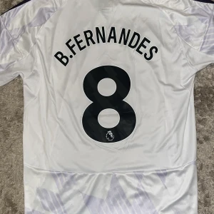 Manchester United B.Fernandes tröja L - Snygg Manchester United fotbollströja med B.Fernandes och nummer 8 på ryggen. Tröjan är vit med ljusa mönster, svarta detaljer och Adidas-logga. Klassiska tre ränder på axlarna och Snapdragon sponsortryck på bröstet. Slim fit-modell i lätt och ventilerande material.