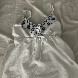 Vit blus/linne med blå broderi - Superfin vit blus/linne med tunna axelband och blått blommigt broderi vid bysten. Den har knyt under bysten