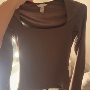 Brun långärmad topp med fyrkantig hals - Säljer en stilren brun långärmad tajt topp från H&M med snygg fyrkantig halsringning. Toppen har en slim passform och är gjord i mjuk bomull som sitter skönt mot kroppen. Perfekt att matcha med jeans eller kjol för en clean look.