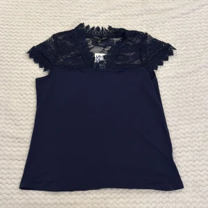 Mörkblå spetsblus från Adrienne Vittadini - Supersnygg mörkblå blus från Adrienne Vittadini med v-ringning och detaljerad spets upptill och på ärmarna. Blusen har korta ärmar och en elegant, feminin look med mjukt tyg nedtill och transparent spets upptill. Perfekt för dig som gillar stilrena och trendiga plagg. (Den har etiketten kvar och är jätte fin på men kommer inte till användning.)