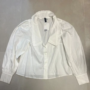 Vit blus med volangkrage från H&M - Säljer en helt ny vit blus från H&M med stor volangkrage och knappar framtill det finns även fläckar. Blusen har långa ärmar med breda manschetter.