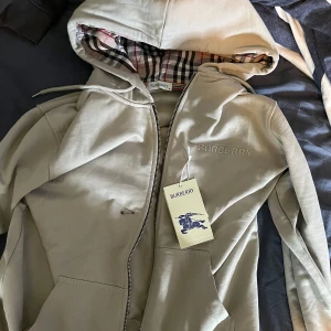 Beige hoodie från Burberry med dragkedja - Säljer en snygg beige hoodie från Burberry. Tröjan har dragkedja, snörning i huvan och broderad Burberry-logga på bröstet. Tillverkad i 100% bomull och har en normal passform. Perfekt för dig som gillar stilrena och exklusiva plagg. Aldrig använd köpt idag.