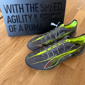 Puma Ultra fotbollsskor grå/gul - Säljer ett par oanvända gråa Puma Ultra fotbollsskor med neon-gula detaljer och snörning. Skorna har en lätt ovandel i syntetmaterial, mönstrad design och dobbar för gräsplan. Insidan är fodrad i gult och det finns en liten svensk flagga på sidan.