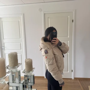 Beige dunbomberjacka Parajumpers S - Säljer en beige dunbomberjacka från Parajumpers med huva, ribbade muddar och flera fickor. Jackan har coola orange detaljer och patch på ärmen. Perfekt för dig som gillar streetstyle och vill ha en varm jacka med snygg passform. Den är använd FÅTAL gånger, köpt på Johnells för 7800.