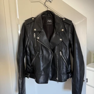 Svart skinnjacka från Zara, M - Snygg svart skinnjacka från Zara med klassisk bikerstil. Jackan har flera silvriga dragkedjor, axelklaffar och bältesdetaljer i midjan. Långärmad modell med coola detaljer och en riktigt edgy vibe. Perfekt för dig som vill ha en tidlös och trendig look!🥰