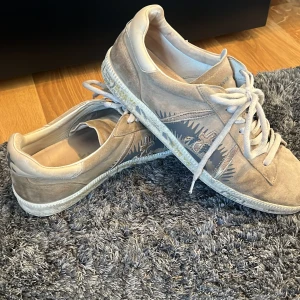 Beige sneakers från Premiata i mocka - Snygga beige sneakers från Premiata i mjuk mocka med coolt grafiskt mönster på sidorna. Skorna har vita snören, rund tå och detaljerad sula med tryck. Perfekta för dig som gillar en unik och stilren look. Skorna och skosnören är lite smutsiga men inget som påbörjar hållbarheten och är lätt att få bort