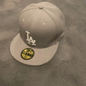 Grå LA-keps från New Era 59FIFTY - Snygg grå keps från New Era 59FIFTY med LA-logga i vitt framtill. Klassisk flat brim och broderade ventilationshål. Passar dig som gillar streetstyle och sportig look. Perfekt för att lyfta din outfit med en ikonisk accessoar.