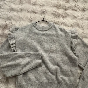 Grå stickad tröja med volang  - Supermysig ljusgrå stickad tröja från Gina Tricot med volangdetaljer längs axlarna. Tröjan har rund halsringning och ribbade muddar. ❣️