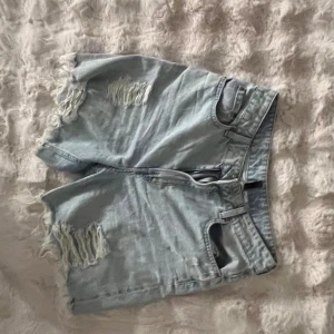 Ljusa jeansshorts från H&M Divided - Snygga ljusblå jeansshorts från H&M Divided i storlek 40. De har slitna detaljer och fransiga kanter för en cool, avslappnad look. Klassisk femficksmodell med dragkedja och knapp. Perfekta för varma dagar och festivaler.
