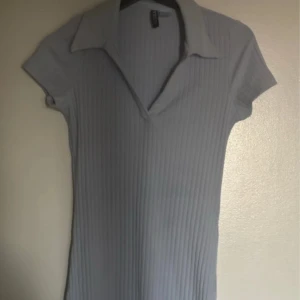 Ljusblå ribbad kortklänning  - Supersöt ljusblå kortklänning  med ribbad struktur, v-ringning och klassisk krage. Klänningen har korta ärmar och en slim passform som framhäver figuren. Perfekt för en trendig och avslappnad look.