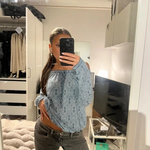 Blå blommig offshoulder blus - Säljer en blå blommig blus med offshoulder och långa ärmar. Toppen har ett tunt, luftigt material och knytband vid halsen. Mönstret är småblommigt i olika blå nyanser och toppen kan bäras både på och utanför axlarna för en chill vibe.