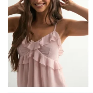 Ljusrosa volangtopp med smala axelband - Supersöt ljusrosa klänning med smala axelband och volangdetaljer över bysten och längs framsidan. Toppen har en luftig passform och är perfekt för varma dagar. Den feminina stilen och de mjuka volangerna ger en romantisk vibe.