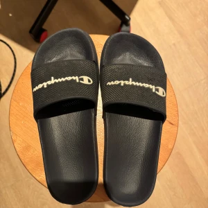 Svarta Champion badsandaler - Mörk blå slides från Champion med vit logga på ovandelen. Skorna har en bred rem i syntetmaterial med meshstruktur och en platt sula. Perfekta att slänga på sig till stranden eller efter träningen. Klassisk och enkel design som funkar till det mesta.