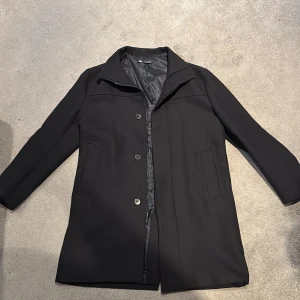 Mörkblå rock från Zara med dragkedja och knapp - Stilren mörkblå rock från Zara med klassisk krage, dold dragkedja och knappar framtill. Jackan har två snedställda fickor och är helfodrad för extra komfort. Perfekt för dig som vill ha en clean och tidlös look under höst och vinter.