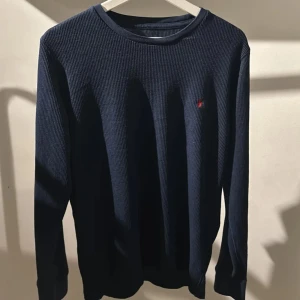 Mörkblå ribbad tröja Polo Ralph Lauren - Mörkblå långärmad tröja från Polo Ralph Lauren med diskret röd logga broderad på bröstet. Tröjan är ribbad och har en klassisk rund halsringning. Perfekt för dig som gillar stilrena och tidlösa plagg.