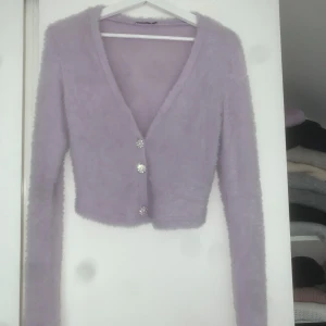 Ljuslila fluffig kofta från Stradivarius - Superfin ljuslila fluffig kofta från Stradivarius med glittriga knappar framtill. Koftan är croppad, har lång ärm och v-ringning. Passar även S