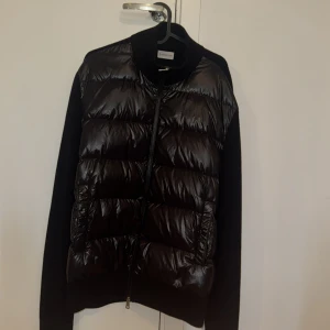  Svart dunväst/jacka från Moncler - Äkta Svart dunväst från Moncler med quiltad framsida i glansig polyester och stickade ärmar samt rygg. Västen har ståkrage, två fickor och Moncler-logga på vänster ärm. Perfekt för dig som gillar stilren och sportig look.
