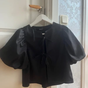 Svart blus med puffärm från Gina Tricot - Svart blus från Gina Tricot i storlek XS med korta puffärmar och två knytningar framtill. Modellen är croppad och har en rak passform. Perfekt för dig som gillar trendiga detaljer och en stilren look.💓💓