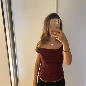 Vinröd offshoulder topp - Säljer en vinröd offshoulder topp med tight passform. Toppen har en snygg drapering och är figurnära, perfekt för dig som gillar en stilren och trendig look. Materialet är mjukt och stretchigt, vilket gör den bekväm att bära.