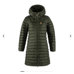 Grön lång dunjacka från Fjällräven - Snygg lång dunjacka från Fjällräven i mörkgrön färg. Jackan har huva, dragkedja hela vägen, diskret logga på bröstet och quiltad design. Perfekt för kalla dagar och riktigt skön att bära. Kommer med tillhörande packpåse.