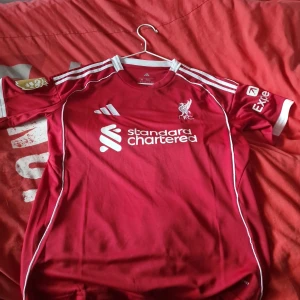 Liverpool matchtröja Adidas röd Wirtz 7 - Liverpool FC matchtröja i rött från Adidas med vita detaljer och tryck. Nummer 7 och WIRTZ på ryggen, Standard Chartered som sponsor framtill. Klassisk design med vita ränder på axlarna och klubbmärke på bröstet. Tillverkad i lätt och ventilerande material. Funkar också om man har s 