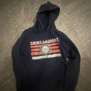 Mörkblå Saint Laurent hoodie - Snygg mörkblå hoodie från Saint Laurent med huva och stor ficka framtill. Framsidan har coolt tryck med röda och vita ränder samt logga. Perfekt för dig som gillar streetwear och vill sticka ut med ett klassiskt märke. Priset kan diskuteras.