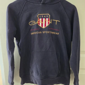Mörkblå hoodie från GANT - Snygg mörkblå hoodie från GANT med broderad logga och sköld på bröstet. Tröjan har huva, magficka och långa ärmar. Tillverkad i mjuk bomullsmix som är skön att ha på sig. Perfekt för dig som gillar klassisk amerikansk stil.