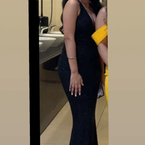 Svart glittrig långklänning Ohpolly  - Säljer en svart långklänning med glittriga detaljer och figurnära passform. Klänningen har en snygg omlottdrapering vid bysten och är axelbandslös, vilket ger en elegant look. Perfekt för bal eller festliga tillfällen när du vill sticka ut.