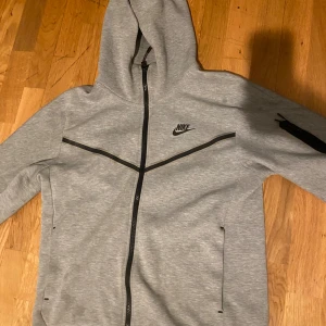 Grå Nike Tech Fleece hoodie S - Nike Tech Fleece hoodie i ljusgrått med svarta detaljer och dragkedja. Tröjan har huva, hel dragkedja, fickor på sidorna och en extra ficka med dragkedja på ärmen. Perfekt för chill eller träning, klassisk Nike-logga på bröstet.
