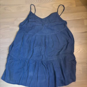 Blå sommarklänning från Green Folk - Luftig blå kortklänning från Green Folk med tunna axelband och v-ringad hals. Klänningen har en avslappnad passform och är perfekt för varma dagar och enkel att styla med sneakers eller sandaler.