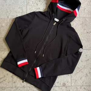 Svart Moncler ziphoodie med logga - Svart hoodie från Moncler med hel dragkedja, huva och snörning. Ribbade muddar med röd, vit och blå detalj samt Moncler-logga på ärmen. Storlek S och toppen skick, fraktas inom 24h✅