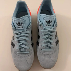  Adidas Gazelle blå   sneakers - Säljer ett par Adidas Gazelle sneakers i ljusblå mocka med svarta klassiska stripes och gummisula. Skorna har snörning, rund tå och en snygg orange detalj på hälen. Perfekta för dig som gillar retrostil och vill sticka ut med färg.