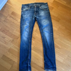 Nudie Grim tim W33 - Snygga blå jeans från Nudie Jeans med klassisk femficksdesign och orangea sömmar. Jeansen har slitningar och en cool tvättad look, samt Nudie-logga på bakfickorna. Lagat hål på låret som ger lite extra charm!