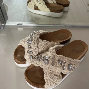 Beige sandaler med flätad ovandel och stenar - Snygga sandaler från Clou med flätad beige ovandel i bomull och stora glittrande stenar. De har en vit plattformsula och korkdetaljer, samt öppen tå för en chill sommarlook. Perfekta att glida runt i när det är varmt ute.