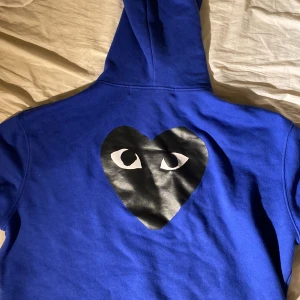 Blå Comme des Garçons PLAY hoodie - Säljer en blå hoodie från Comme des Garçons PLAY med dragkedja och huva. Tröjan har det ikoniska hjärta-med-ögon-trycket i svart och vitt både fram och stort på ryggen. Praktiska fickor och snörning i huvan. Perfekt för dig som gillar streetwear och vill sticka ut.
