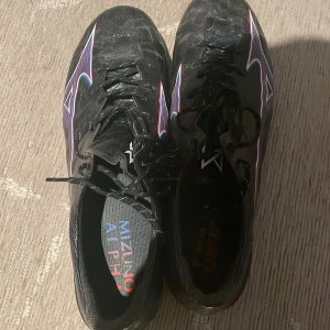 Svarta Mizuno Alpha fotbollsskor - Säljer ett par svarta Mizuno Alpha fotbollsskor med coola mönster i blått, rött och vitt. Skorna har snörning och dobbar för bra grepp på planen. Lätta och smidiga med sportig design, perfekta för dig som vill sticka ut på fotbollsplanen.