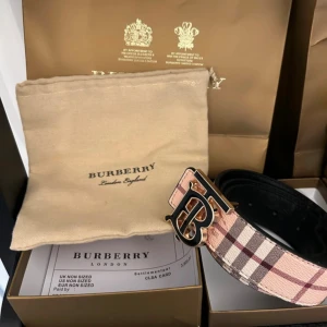  bälte  - Säljer ett snyggt med klassiskt rutig design i beige, brun och rosa toner. Bältet har ett stort metallspänne med Burberrys ikoniska logotyp. Perfekt accessoar för att lyfta din outfit med lyxig känsla och stil.