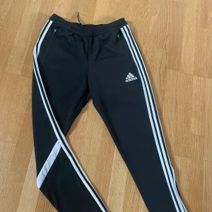 Adidas träningsbyxor  - Svarta träningsbyxor från Adidas med vita ränder längs sidorna och dragkedja vid benslut. Byxorna har elastisk midja med snörning, två fickor med dragkedja och Climacool-material som andas. Perfekta för sport eller chill dagar. Skriv om du har frågor🫶🏼