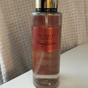 Victoria's Secret Temptation mist - En snygg och fräsch body mist från Victoria's Secret i doften Temptation. Flaskan är genomskinlig med rosa-orange etikett och guldfärgat lock. Rymmer 250 ml och har en feminin, söt och fruktig doft. Perfekt att spraya på kroppen för en fräsch känsla.