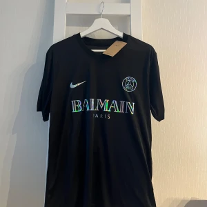 Svart PSG x Balmain fotbollströja Nike - Snygg svart fotbollströja från Nike i samarbete med PSG och Balmain. Tröjan har korta ärmar, rund hals och holografiska detaljer på logga och text. Materialet är mjukt och lätt, perfekt för träning eller match. Ikonisk Paris Saint-Germain badge på bröstet.