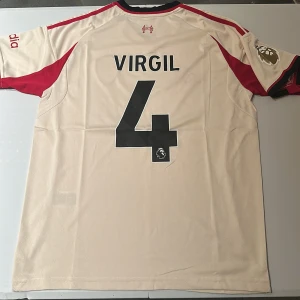 Liverpool Virgil #4 bortatröja Adidas - Liverpool FC bortatröja med Virgil och nummer 4 på ryggen. Beige bas med röda detaljer, Premier League-märke i guld på ärmen och klubbmärke på bröstet. Tillverkad i lätt och ventilerande polyester med AEROREADY-teknologi. Klassisk passform och korta ärmar. Replika, endast handtvätt gäller!