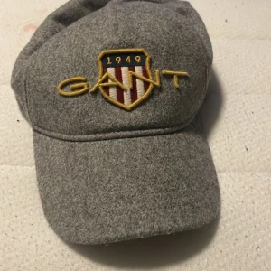 Grå ullkeps från GANT med brodyr - Snygg grå keps från GANT i ullmaterial med broderad GANT-logga och sköld framtill. Justerbar rem i brunt skinn baktill med metallspänne. Perfekt accessoar för dig som vill ha en klassisk och stilren look.