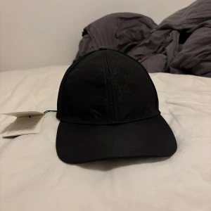 Svart keps från Gucci med meshdetaljer - Snygg svart keps från Gucci med diskret GG-monogram över hela framsidan. Kepsen har mesh på baksidan för extra ventilation och justerbar snapback. Tillverkad i polyester och har en stilren, sportig look med märkets logga på insidan och etikett bak. Kepsen är helt ny och aldrig användits, fick den som present men den passar inte mig. Har även dustbagen till kepsen.