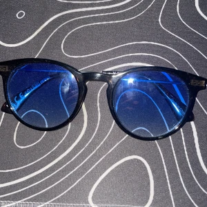 Svarta runda glasögon med blått glas - Snygga svarta glasögon med rund form och blåtonade glas. Bågarna är i plast och har en klassisk look som passar till många stilar. Perfekta för dig som vill sticka ut med en cool detalj. Metallgångjärn för extra stabilitet.